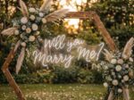 'Will You Marry Me?' neon sign displayed beneath a boho wooden arch with white pampas grass and floral arrangements in an outdoor garden setting