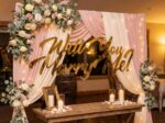 Gold cursive 'Will You Marry Me?' neon sign glowing against a romantic blush curtain backdrop with candles and white floral arch inside an intimate venue