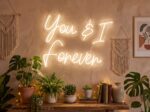 'You & I Forever' warm white neon sign mounted on a wall in a cosy boho-styled room with plants and macramé decor