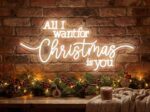 'All I Want for Christmas Is You' white neon sign glowing against a festive brick wall with holiday greenery and warm string lights