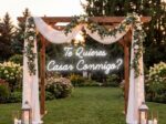 'Te Quieres Casar Conmigo?' Spanish 'Will You Marry Me' neon sign under a floral archway with lanterns in an outdoor garden for a bilingual proposal