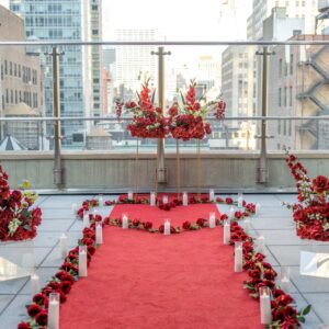 Red carpet proposal pathway lined with tall red floral stands, rose petals and candles on a rooftop terrace with Midtown Manhattan skyline views
