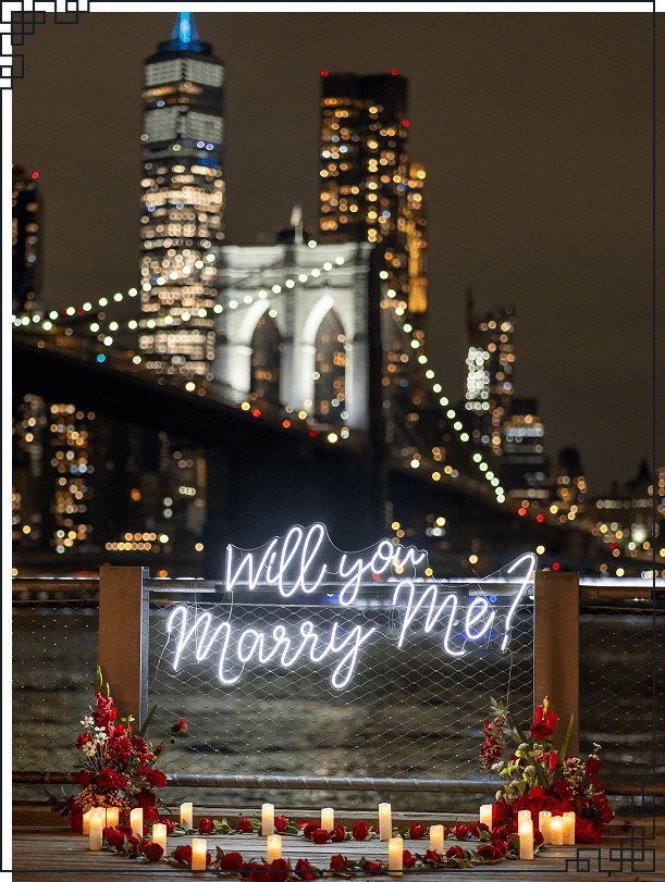 Nighttime NYC proposal with 'Will You Marry Me' neon sign and rose petals near the Brooklyn Bridge with city lights reflected on the water