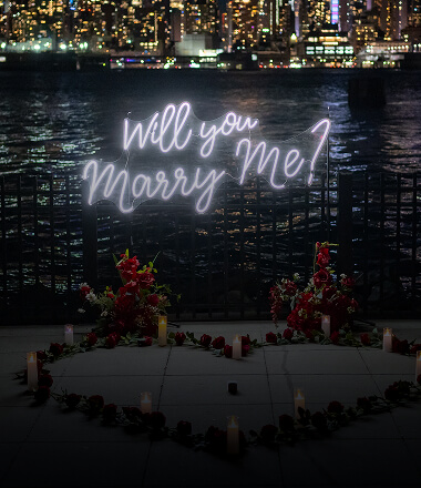 Glowing 'Will You Marry Me' neon sign surrounded by rose petals and candles in a heart shape with the lit-up New York City skyline at night