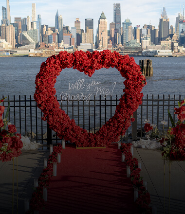 Luxury red rose heart archway proposal setup with red carpet and floral arrangements on a waterfront pier overlooking the Manhattan skyline