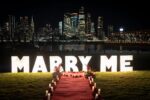 Illuminated MARRY ME marquee letter signs on green lawn with red carpet pathway lined with candles and rose arrangements against a sparkling NYC night skyline