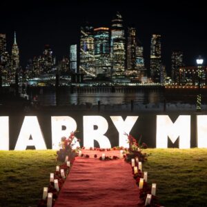 Illuminated MARRY ME marquee letter signs on green lawn with red carpet pathway lined with candles and rose arrangements against a sparkling NYC night skyline
