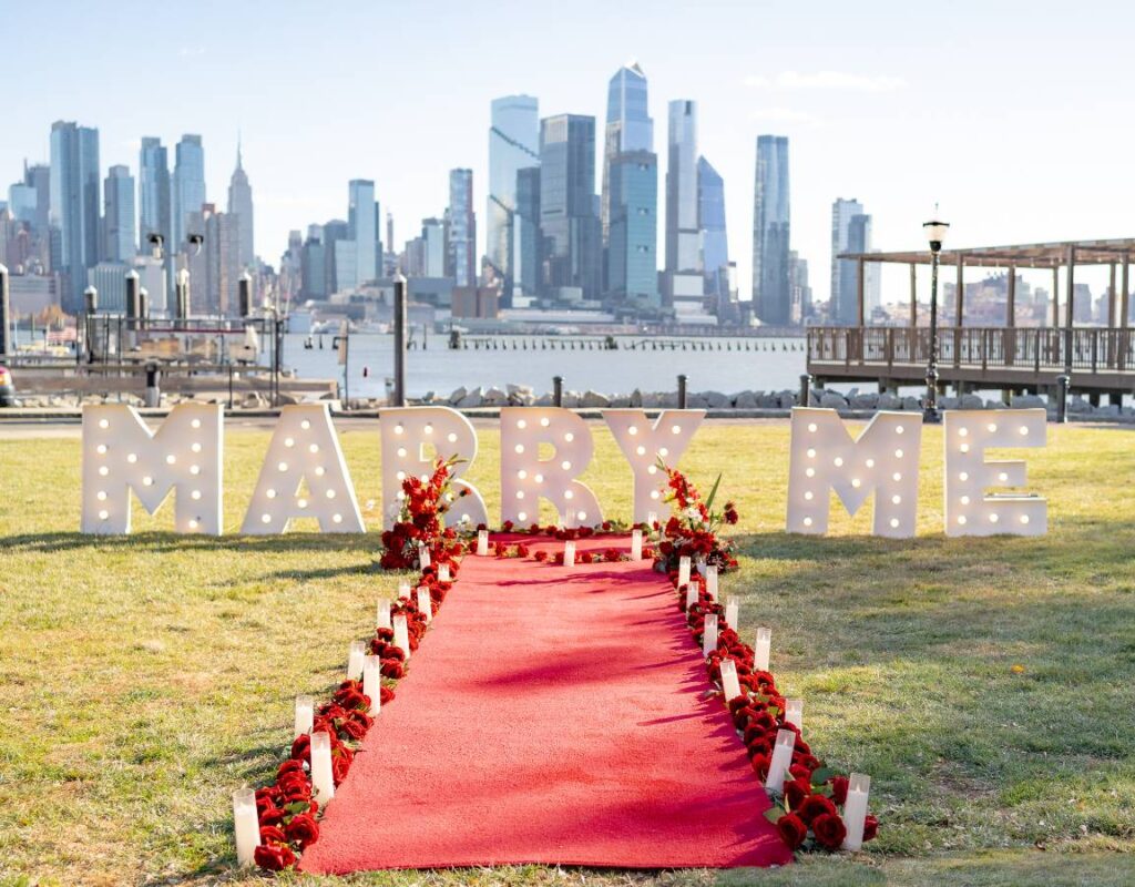 Daytime MARRY ME marquee letters on waterfront lawn with red carpet and red floral arrangements, Lower Manhattan skyline and Hudson River in the background