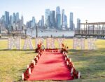 Daytime MARRY ME marquee letters on waterfront lawn with red carpet and red floral arrangements, Lower Manhattan skyline and Hudson River in the background