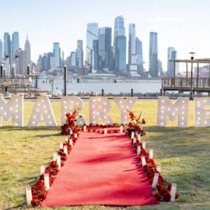 Daytime MARRY ME marquee letters on waterfront lawn with red carpet and red floral arrangements, Lower Manhattan skyline and Hudson River in the background