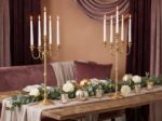 Gold five-arm candelabra with white taper candles on a wooden table with white roses and greenery garland for an elegant romantic proposal setting