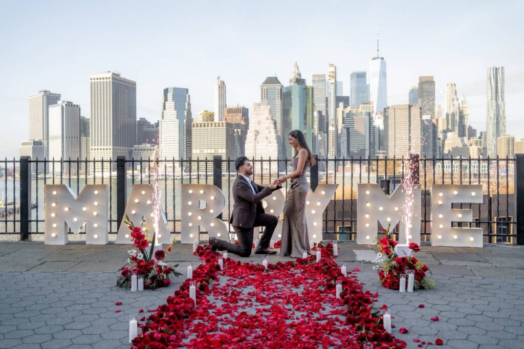 Man proposing on one knee amid a carpet of red rose petals beside MARRY ME marquee letters on a waterfront rooftop with the Lower Manhattan skyline
