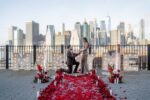 Man proposing on one knee amid a carpet of red rose petals beside MARRY ME marquee letters on a waterfront rooftop with the Lower Manhattan skyline