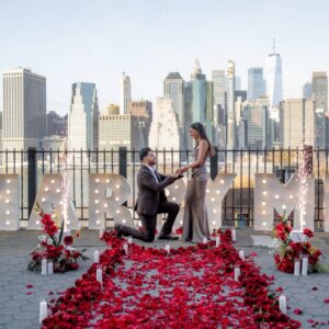 Man proposing on one knee amid a carpet of red rose petals beside MARRY ME marquee letters on a waterfront rooftop with the Lower Manhattan skyline