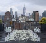 White rose petal pathway with floral arrangements and 'You & I Forever' sign on a rooftop terrace overlooking Lower Manhattan and the Hudson River