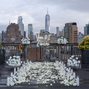 White rose petal pathway with floral arrangements and 'You & I Forever' sign on a rooftop terrace overlooking Lower Manhattan and the Hudson River