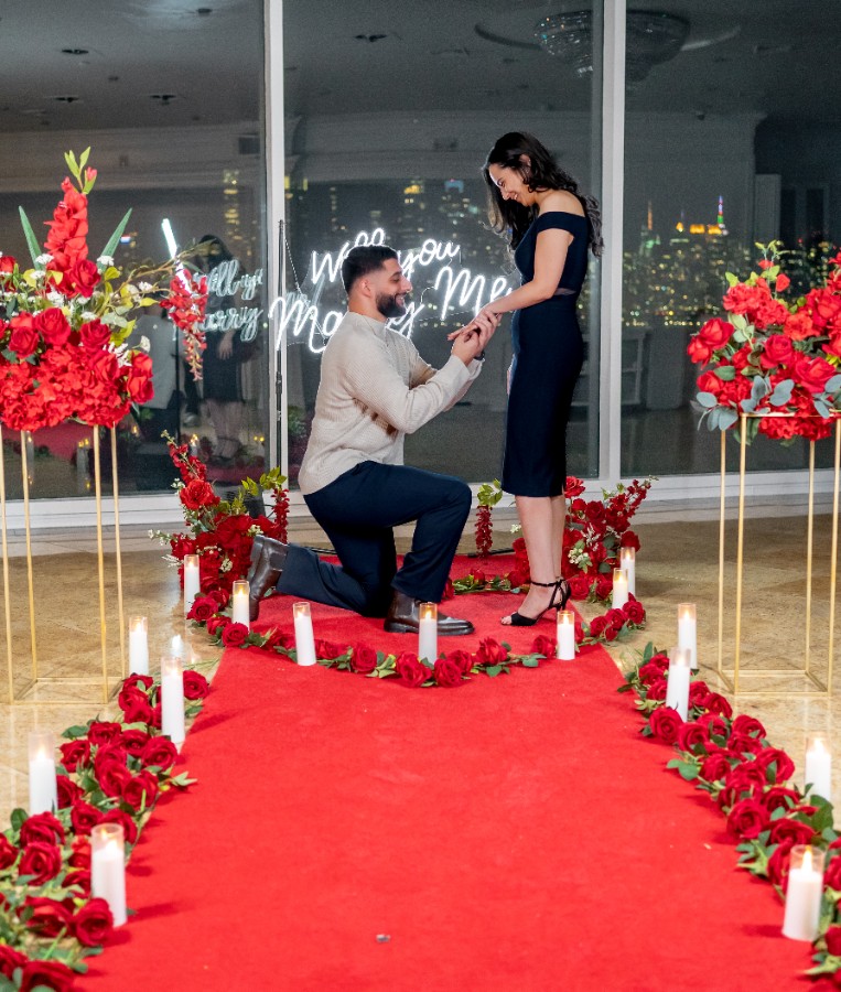 Man proposing on one knee along a red carpet lined with LED candles and red roses inside a bright high-rise venue with 'Will You Marry Me' neon sign visible