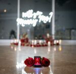 'Will You Marry Me' neon sign glowing above a heart of red silk roses with a ring box on the floor of a high-rise venue with city views