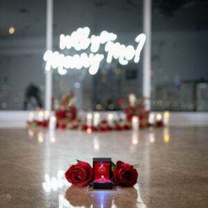 'Will You Marry Me' neon sign glowing above a heart of red silk roses with a ring box on the floor of a high-rise venue with city views