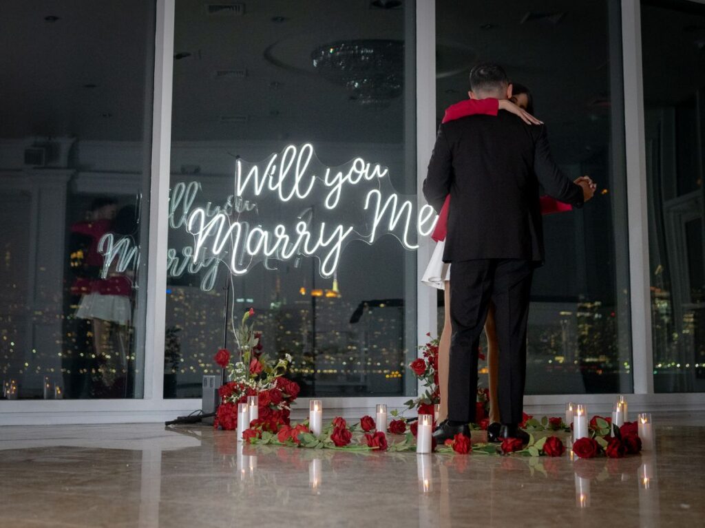 Man proposing to his partner in front of a 'Will You Marry Me' neon sign with silk roses and candles, NYC skyline visible through floor-to-ceiling windows at night