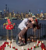 Couple sharing a romantic kiss beside red rose floral stands and candles at night with the glittering New York City skyline as a backdrop