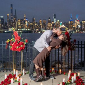 Couple sharing a romantic kiss beside red rose floral stands and candles at night with the glittering New York City skyline as a backdrop