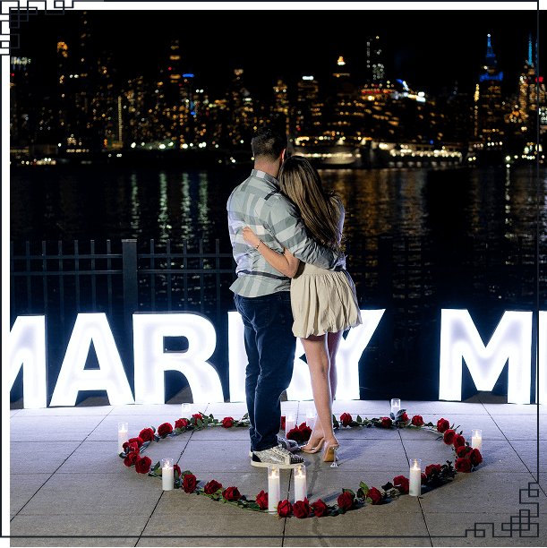 Couple embracing beside large illuminated MARRY ME letter signs on a waterfront promenade with the glittering NYC skyline in the background