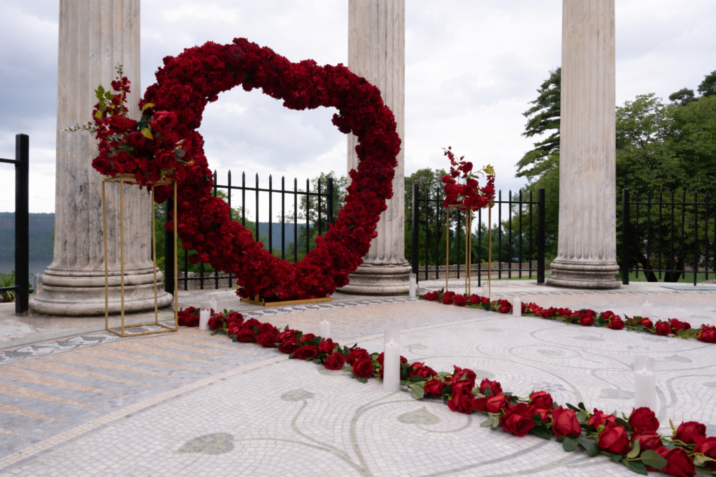 Red rose heart arch rental setup at a stone colonnade outdoor venue with red rose petals scattered on the ground for a romantic proposal