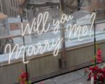 'Will You Marry Me?' neon sign mounted on mounting poles with pink floral arrangement against glass windows and NYC rooftop view