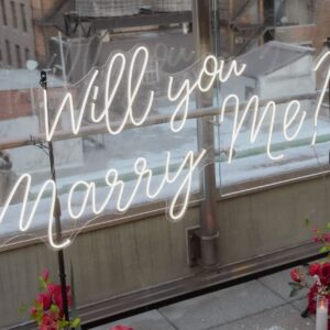 'Will You Marry Me?' neon sign mounted on mounting poles with pink floral arrangement against glass windows and NYC rooftop view