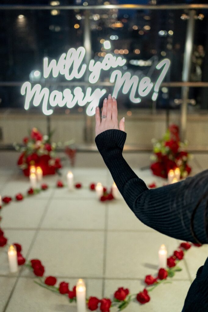 Woman reaching up to touch a glowing 'Will You Marry Me?' neon sign surrounded by rose petals and candles in a romantic proposal setting