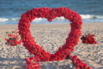 Red rose heart arch on a sandy beach with ocean waves in the background for a romantic seaside marriage proposal