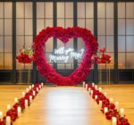 Stunning red rose heart-shaped arch with 'Will You Marry Me' calligraphy sign in a grand indoor hall, flanked by candle-lined aisle and floor-to-ceiling windows