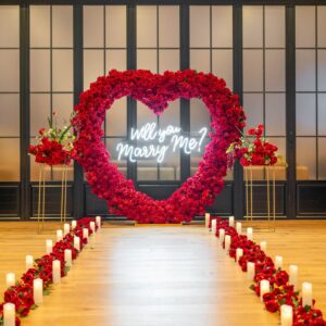 Stunning red rose heart-shaped arch with 'Will You Marry Me' calligraphy sign in a grand indoor hall, flanked by candle-lined aisle and floor-to-ceiling windows