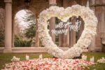 White floral heart arch inscribed with 'Will You Marry Me' framing a garden colonnade pathway covered in rose petals and candles