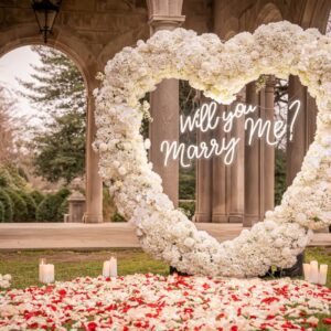 White floral heart arch inscribed with 'Will You Marry Me' framing a garden colonnade pathway covered in rose petals and candles
