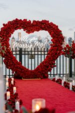 Red rose heart archway with 'Will You Marry Me' sign set against wrought iron gates with red carpet and candles at dusk