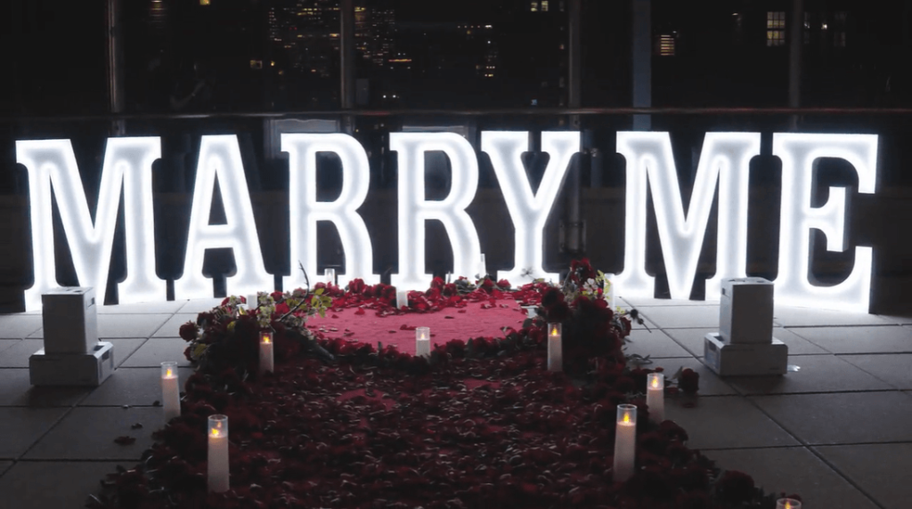 Close-up night view of glowing MARRY ME marquee letters surrounded by rose petals and candles inside a dimly lit elegant venue