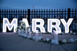 MARRY ME large letter signs with white floral arrangements at night with city fence and soft blue evening sky in the background