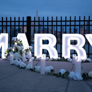 MARRY ME large letter signs with white floral arrangements at night with city fence and soft blue evening sky in the background