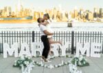 Couple embracing beside MARRY ME letter signs with white roses and floral garlands, Manhattan skyline visible across the river