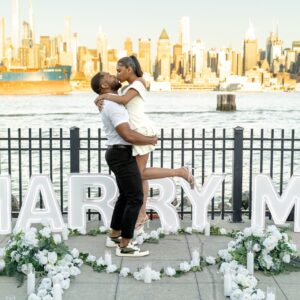 Couple embracing beside MARRY ME letter signs with white roses and floral garlands, Manhattan skyline visible across the river