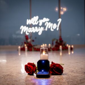 Close-up of 'Will You Marry Me' neon sign glowing in blue light surrounded by red roses and candles in an intimate indoor setting