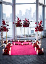 Romantic indoor proposal setup with red floral arrangements and pink rose petal pathway leading to floor-to-ceiling windows with the New York City skyline