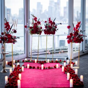 Romantic indoor proposal setup with red floral arrangements and pink rose petal pathway leading to floor-to-ceiling windows with the New York City skyline
