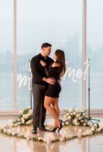 Couple standing beside a 'Will You Marry Me' neon sign with white floral semicircle arrangement and NYC skyline through floor-to-ceiling windows