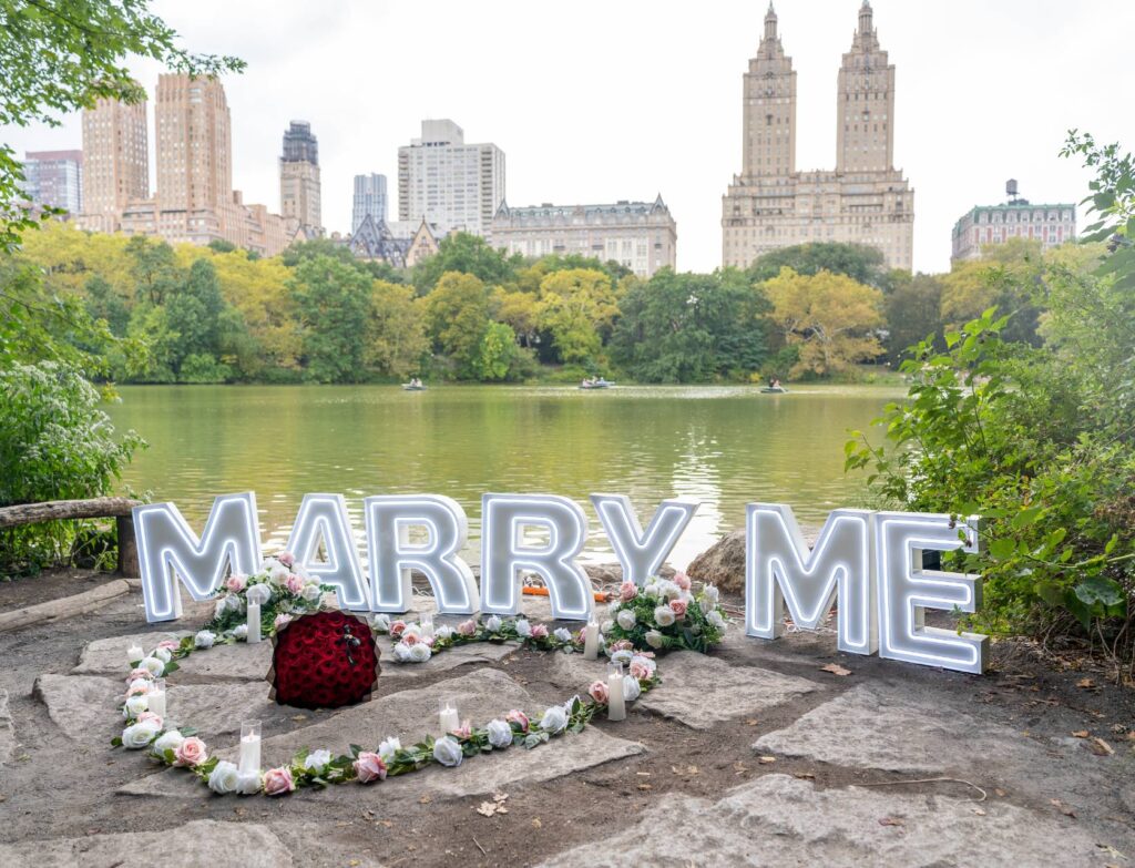 MARRY ME letter signs surrounded by white floral wreaths beside The Lake in Central Park with iconic Upper West Side buildings in the background