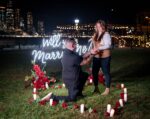 Man proposing on one knee in a candlelit outdoor setting with 'Will You Marry Me' neon sign and red roses, NYC lights glowing in the background