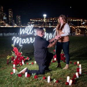 Man proposing on one knee in a candlelit outdoor setting with 'Will You Marry Me' neon sign and red roses, NYC lights glowing in the background