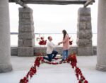 Man down on one knee proposing to his partner beneath a stone pergola with a red rose petal pathway, NYC waterfront in the background